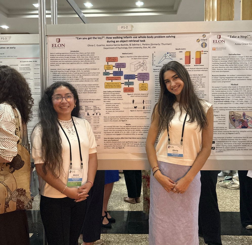Two students stand in front of a research poster at an academic conference, smiling at the camera. Both wear conference name badges as attendees and other research posters fill the background.